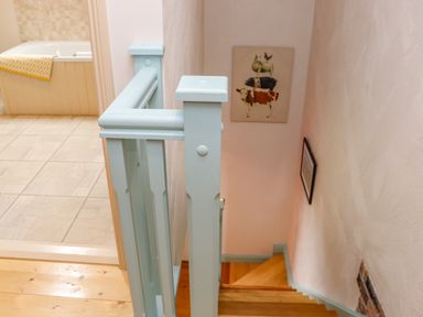 A staircase with light blue painted railing and wooden steps next to a bathroom with tiled floor and a bathtub with a yellow towel at Cob Cottage in Kilmore County Wexford