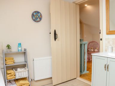 A bathroom with a shelf holding yellow towels and a plant near an open door showing a pink chair with teddy bears at Cob Cottage in Kilmore County Wexford