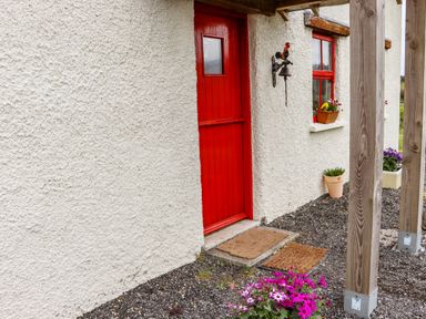 A red door on a white textured wall with flowers in pots and a rooster bell at Cob Cottage in Kilmore County Wexford