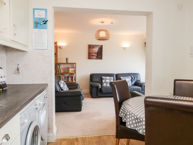 A kitchen area with washing machine and countertop next to an open doorway leading to a living room with black sofas a bookshelf a painting and a dining table with chairs at Rothay in Ambleside