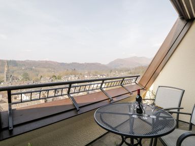 A balcony with a round table holding a bottle of wine and four wine glasses with two chairs and a view of buildings and hills at Rothay in Ambleside