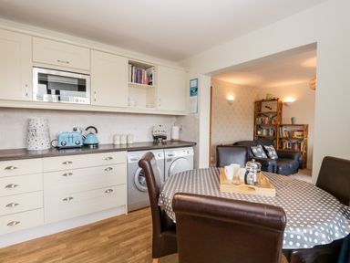 A kitchen with white cabinets a countertop with toaster kettle and canisters and a dining table with chairs next to a living room with bookshelves and sofas at Rothay in Ambleside