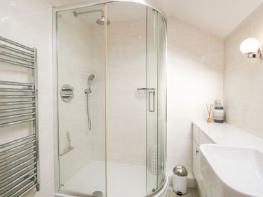 A bathroom with a curved glass shower enclosure a wall-mounted towel heater a white sink and countertop with cabinets and bathroom accessories at Rothay in Ambleside
