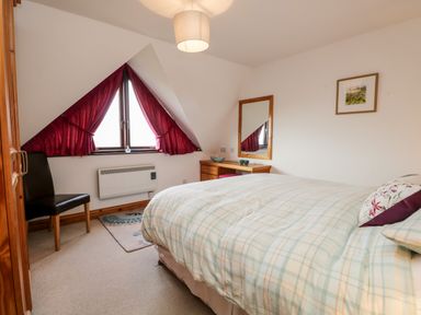 A bedroom with a bed a wooden desk with a mirror a black chair and a triangular window with red curtains at Rothay in Ambleside