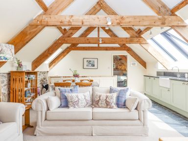 An open living space with wooden beams a beige sofa with cushions a dining table and a kitchen area with light green cabinets at The Coach House at Thorn Farm Bridford near Moretonhampstead