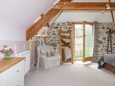 A bedroom with an armchair a floor lamp a standing mirror an open door and a dresser with flowers at The Coach House at Thorn Farm Bridford near Moretonhampstead