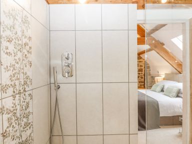 A tiled shower area with floral patterned tiles on one wall and a glass door showing a bedroom with bed pillows and exposed wooden beams at The Coach House at Thorn Farm Bridford near Moretonhampstead