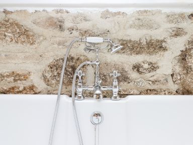 A bathtub with a chrome faucet and handheld showerhead against a stone wall at The Coach House at Thorn Farm Bridford near Moretonhampstead