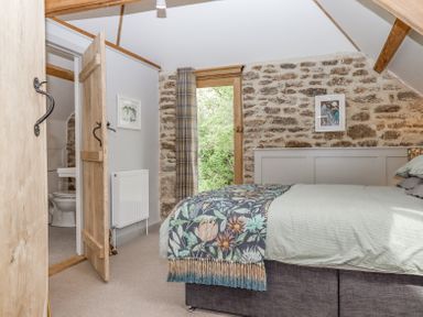 A bedroom with a bed and floral blanket a stone wall a door leading outside and a door to a bathroom at The Coach House at Thorn Farm Bridford near Moretonhampstead