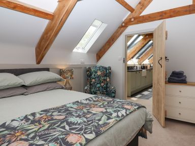 A bedroom with a bed a floral armchair a lamp and wooden beams with a view into a kitchen at The Coach House at Thorn Farm Bridford near Moretonhampstead