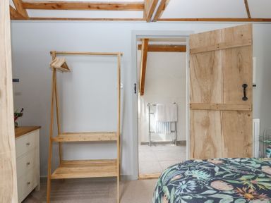 A bedroom with a wooden clothes rack, a chest of drawers, a floral bedspread, and an open wooden door leading to a bathroom with a towel rail at The Coach House at Thorn Farm Bridford near Moretonhampstead