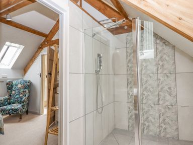 A bathroom with a glass shower enclosure and patterned tiles next to a room with a floral armchair at The Coach House at Thorn Farm in Bridford near Moretonhampstead