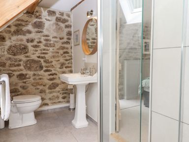A bathroom with a toilet pedestal sink round mirror stone wall and glass shower door at The Coach House at Thorn Farm in Bridford near Moretonhampstead