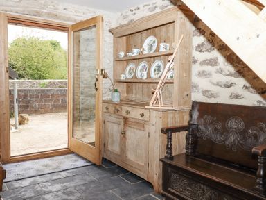 A wooden cupboard with plates and cups next to a dark wooden bench under a staircase near an open glass door at The Coach House at Thorn Farm Bridford near Moretonhampstead