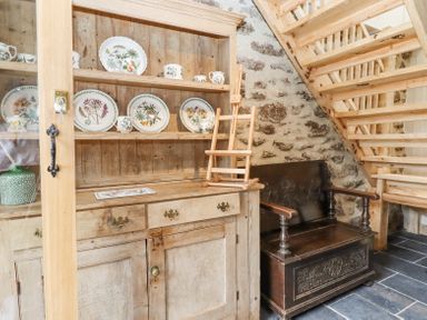 A wooden cabinet with plates and mugs next to a dark wooden bench under wooden stairs with a stone wall in the background at The Coach House at Thorn Farm Bridford near Moretonhampstead