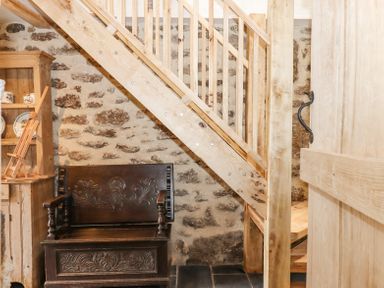 A wooden staircase over a carved wooden bench and a wooden cabinet with dishes against a stone wall at The Coach House at Thorn Farm Bridford near Moretonhampstead