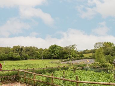 A fenced green field with trees and a gravel area with obstacles in the distance at The Coach House at Thorn Farm in Bridford near Moretonhampstead