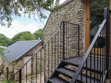 An outdoor metal staircase leading to a glass door on a stone building with another stone building and trees in the background at The Coach House at Thorn Farm Bridford near Moretonhampstead