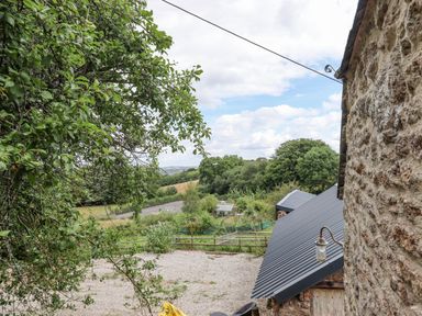 A stone building with a slanted metal roof beside trees and a fenced garden area at The Coach House at Thorn Farm in Bridford near Moretonhampstead