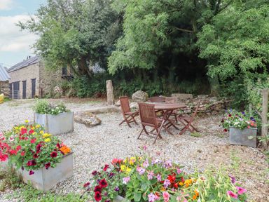 An outdoor gravel area with wooden table and chairs surrounded by flower planters and trees at The Coach House at Thorn Farm Bridford near Moretonhampstead
