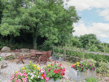An outdoor seating area with wooden chairs and table surrounded by flower planters and trees at The Coach House at Thorn Farm in Bridford near Moretonhampstead