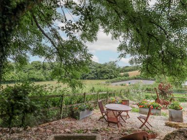 An outdoor seating area with a round table and four wooden chairs surrounded by flower planters and trees at The Coach House at Thorn Farm Bridford near Moretonhampstead