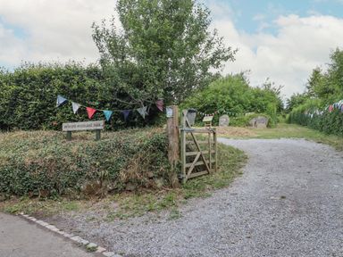 A gravel path leading to a wooden gate and green hedge with a sign reading Bridford Woodland Park with bunting strung across the area at The Coach House at Thorn Farm Bridford near Moretonhampstead