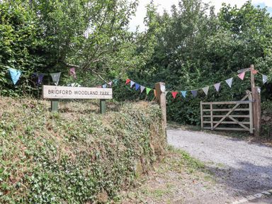 A gated entrance with colorful pennant flags and a sign reading Bridford Woodland Park surrounded by greenery at The Coach House at Thorn Farm Bridford near Moretonhampstead