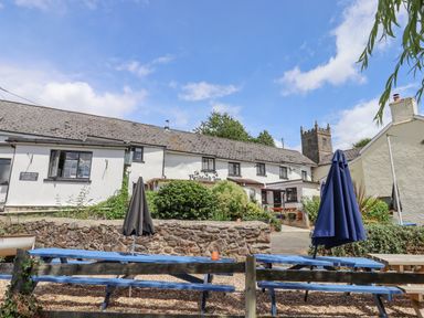 Outdoor seating with blue picnic tables and closed umbrellas in front of a white building with a stone wall and a church tower in the background at The Coach House at Thorn Farm in Bridford near Moretonhampstead