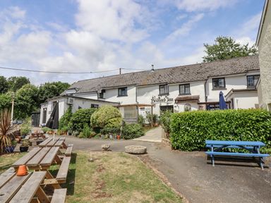 An outdoor seating area with wooden picnic tables and umbrellas in front of a white building with shrubbery at The Coach House at Thorn Farm in Bridford near Moretonhampstead