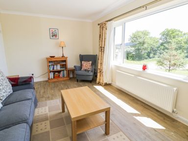 A living room with a wooden coffee table a blue sofa a blue armchair beside a bookshelf and a large window showing trees at Ceris Penrhyndeudraeth