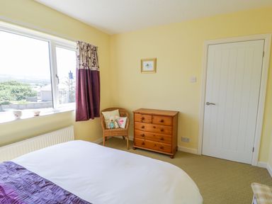 A bedroom with a bed a wooden dresser a wicker chair with cushions a window and a white door at Ceris Penrhyndeudraeth