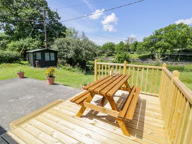 A wooden picnic table on a wooden deck overlooking a grassy garden with trees and a black shed at Ceris in Penrhyndeudraeth