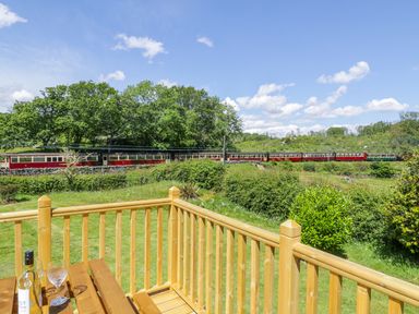 A wooden deck with a table holding a bottle and glasses overlooking a grassy area with bushes and a train passing in the background at Ceris in Penrhyndeudraeth