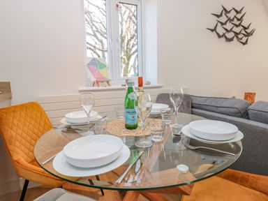 A dining area with a round glass table set with plates glasses cutlery and bottles near a window and grey sofa at Seaspray in Stoke Fleming Near Dartmouth