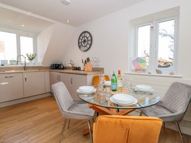 A dining area with a glass round table set for four and a kitchen with cabinets and windows at Seaspray in Stoke Fleming Near Dartmouth