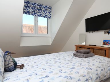 A bedroom with a bed covered in patterned bedding a wooden dresser with baskets a mounted television and a window with patterned curtains at Seaspray Stoke Fleming Near Dartmouth