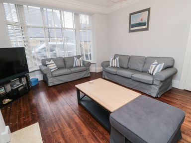A living room with two gray sofas, a wooden coffee table, a large window with blinds, and a television on a stand at Glenander in Bamburgh