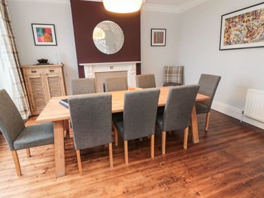 A dining room with a wooden table and eight gray upholstered chairs a wooden cabinet a round mirror above a white fireplace and framed artwork on the walls at Glenander in Bamburgh