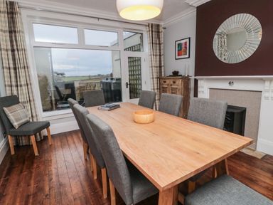 A dining room with a wooden table surrounded by gray chairs a wooden cabinet a fireplace and large windows with plaid curtains at Glenander Bamburgh