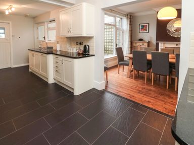 A kitchen with white cabinets and black countertops next to a dining room with a wooden table and gray chairs at Glenander in Bamburgh