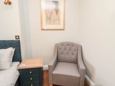 A bedroom corner with a grey armchair next to a bedside table with drawers and a bed with white pillows at Glenander in Bamburgh