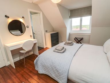 A bedroom with a bed, a desk with a round mirror, a chair, a chest of drawers, and a window at Glenander in Bamburgh