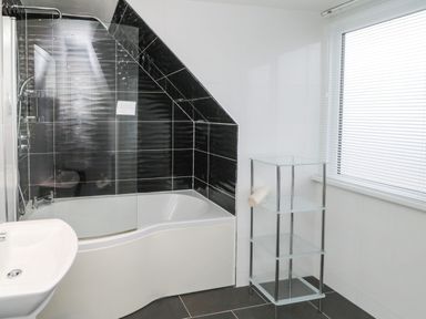 A bathroom with a bathtub and shower area with black tiles a white sink and a glass shelving unit by a window at Glenander in Bamburgh
