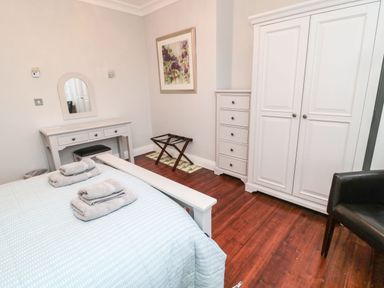 A bedroom with a bed, folded towels, a dressing table with a mirror, a chest of drawers, a wardrobe, a luggage rack, and a chair at Glenander in Bamburgh