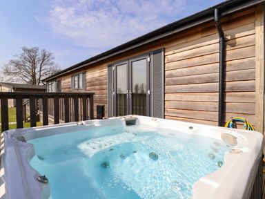 An outdoor hot tub next to a wooden building with glass doors and a wooden railing at Skylarks in Fornham St Genevieve near Bury St Edmunds