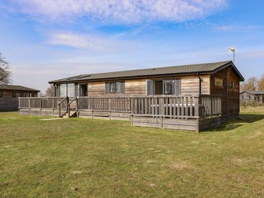 A wooden lodge with a fenced wooden deck on a grassy area at Skylarks in Fornham St Genevieve near Bury St Edmunds