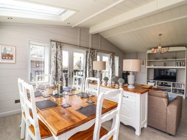 A dining area with a wooden table set with placemats and glassware next to a living room with a TV and shelving at Skylarks in Fornham St Genevieve near Bury St Edmunds