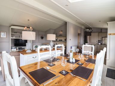 A dining area with a wooden table set with placemats glasses and cutlery in a room with white chairs a kitchen and a living space at Skylarks in Fornham St Genevieve near Bury St Edmunds