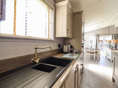 A kitchen with a sink toaster and coffee machine leading to a dining area with white chairs at Skylarks in Fornham St Genevieve near Bury St Edmunds